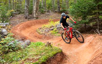 A cyclist riding a red dirt mountain bike trail through a wooded forest.