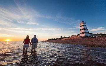 Two people watching the sunset over the water beside a black-and-white striped lighthouse.